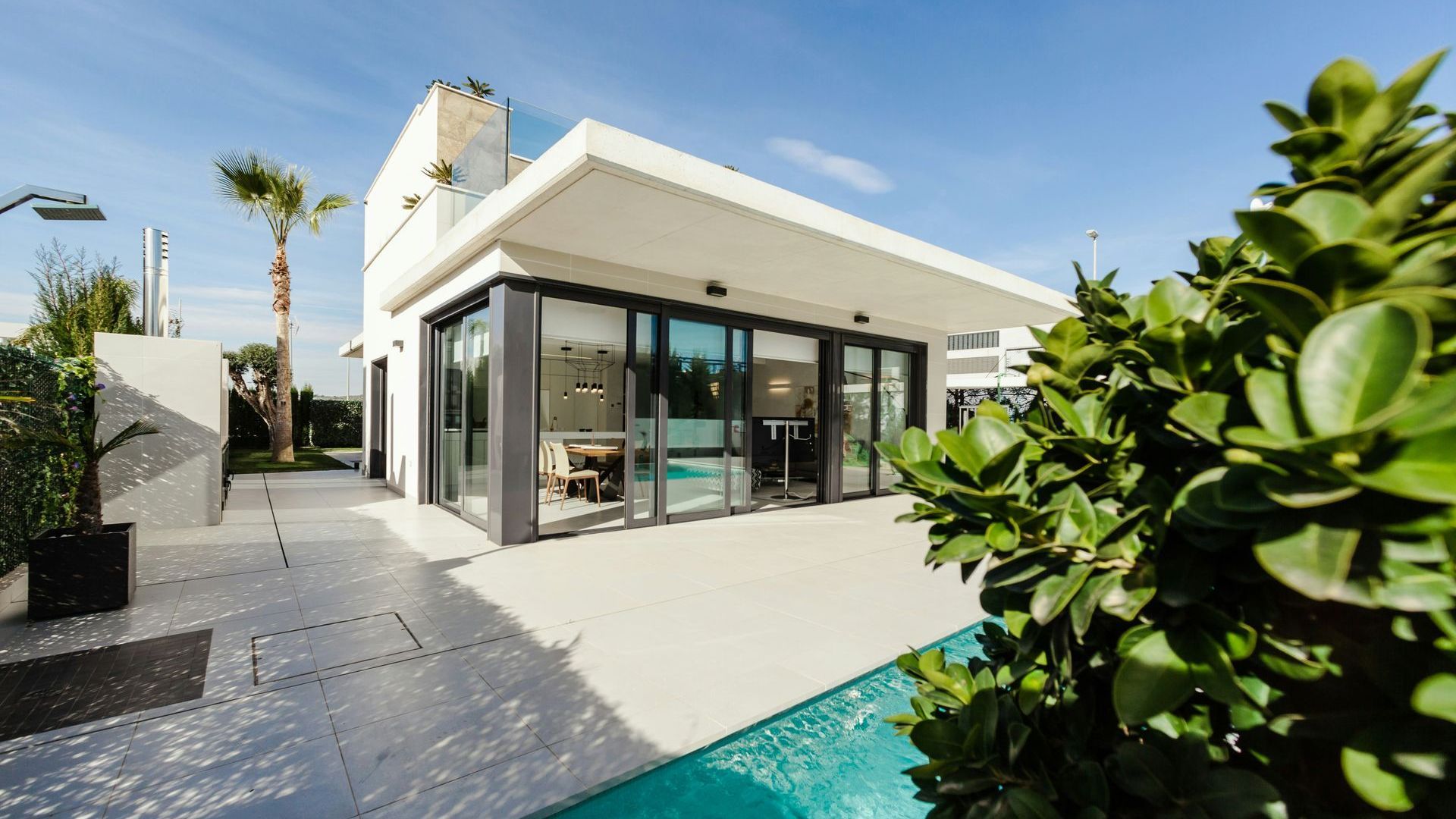 Modern white house with large glass windows beside a turquoise pool and lush greenery.