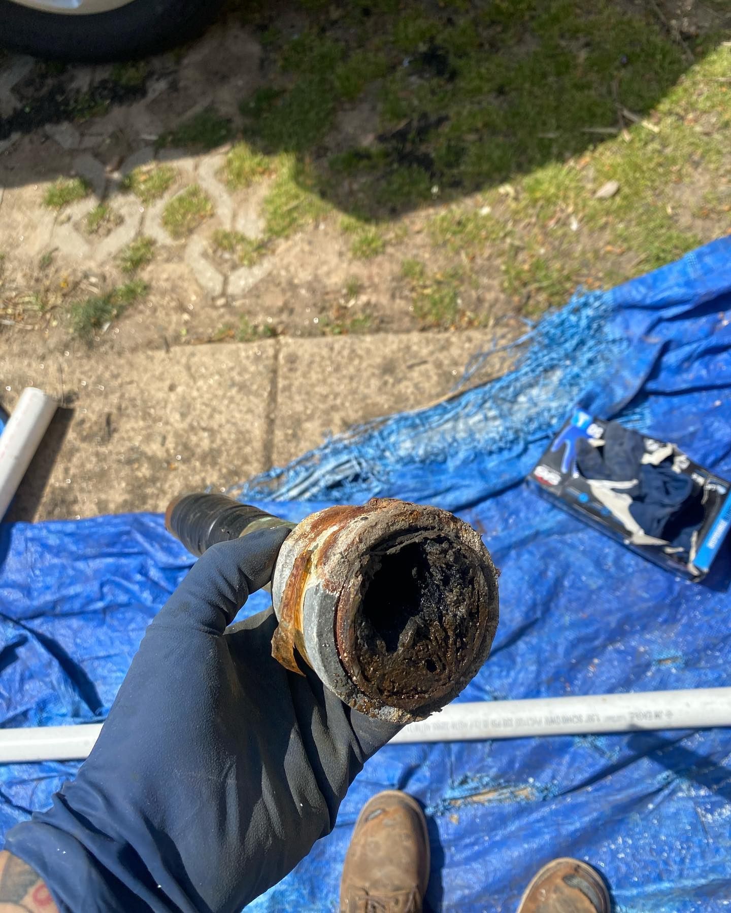 Hand holding a corroded pipe section, showing thick black buildup inside. Outdoors on a blue tarp.
