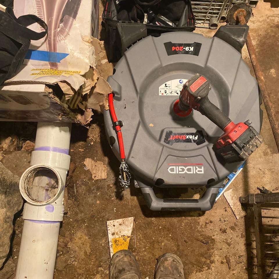 Plumber's tools: sewer snake, drill, and PVC pipe in a basement setting.