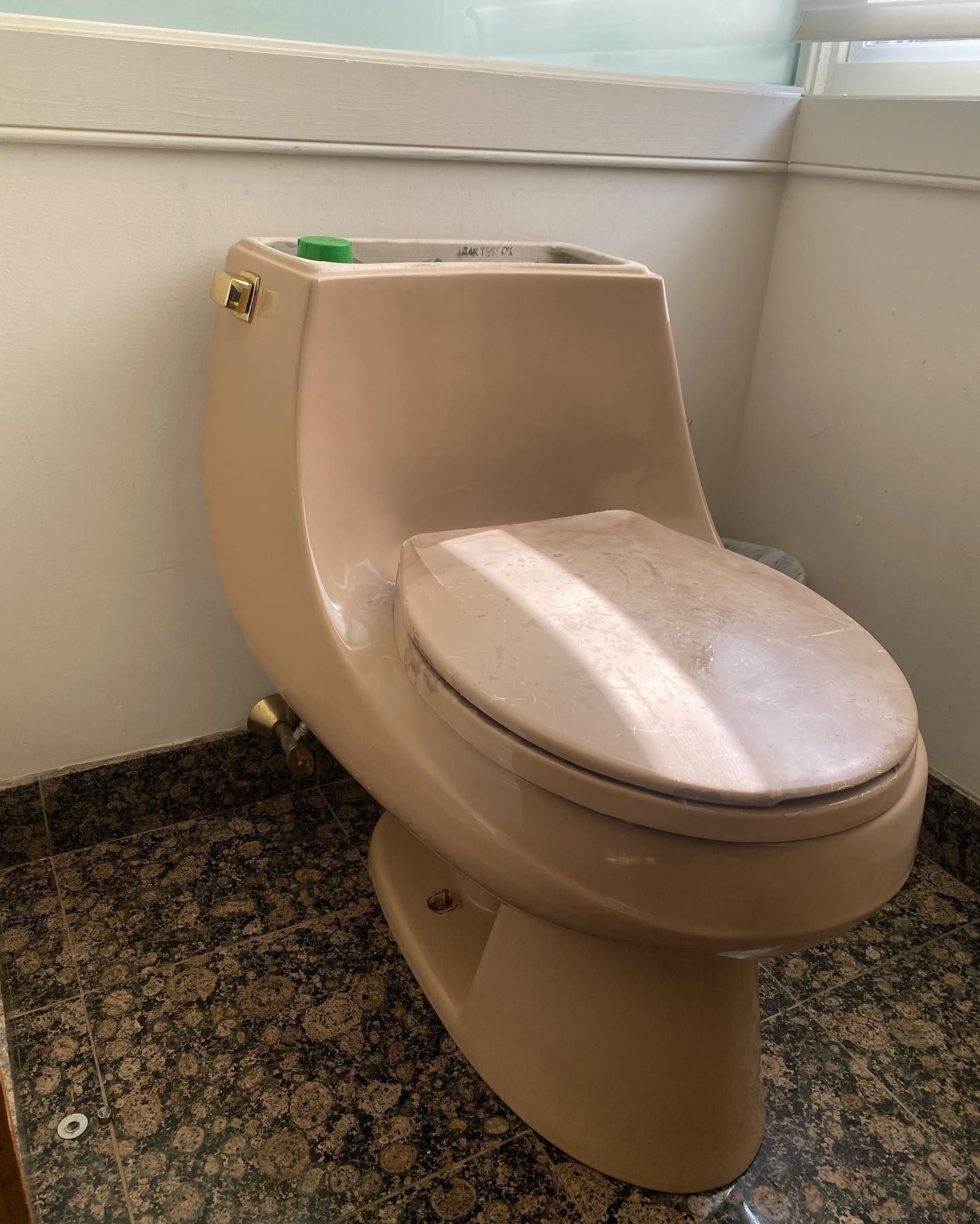 Beige, one-piece toilet with closed lid. Located in a bathroom with a dark speckled floor and white walls.
