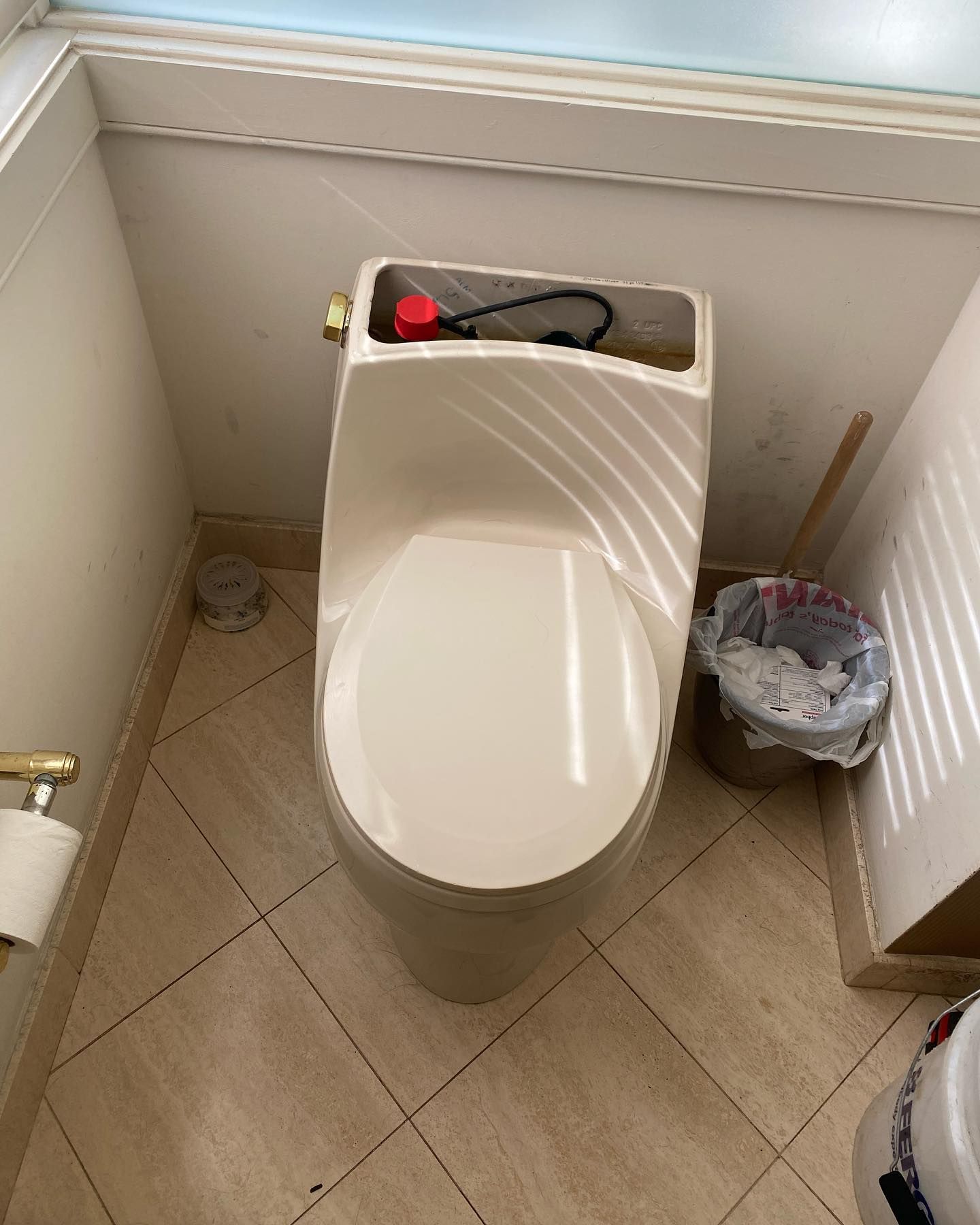 Toilet in a bathroom. Light-colored toilet with seat down, set on tiled floor. Trash can and toilet paper holder present.