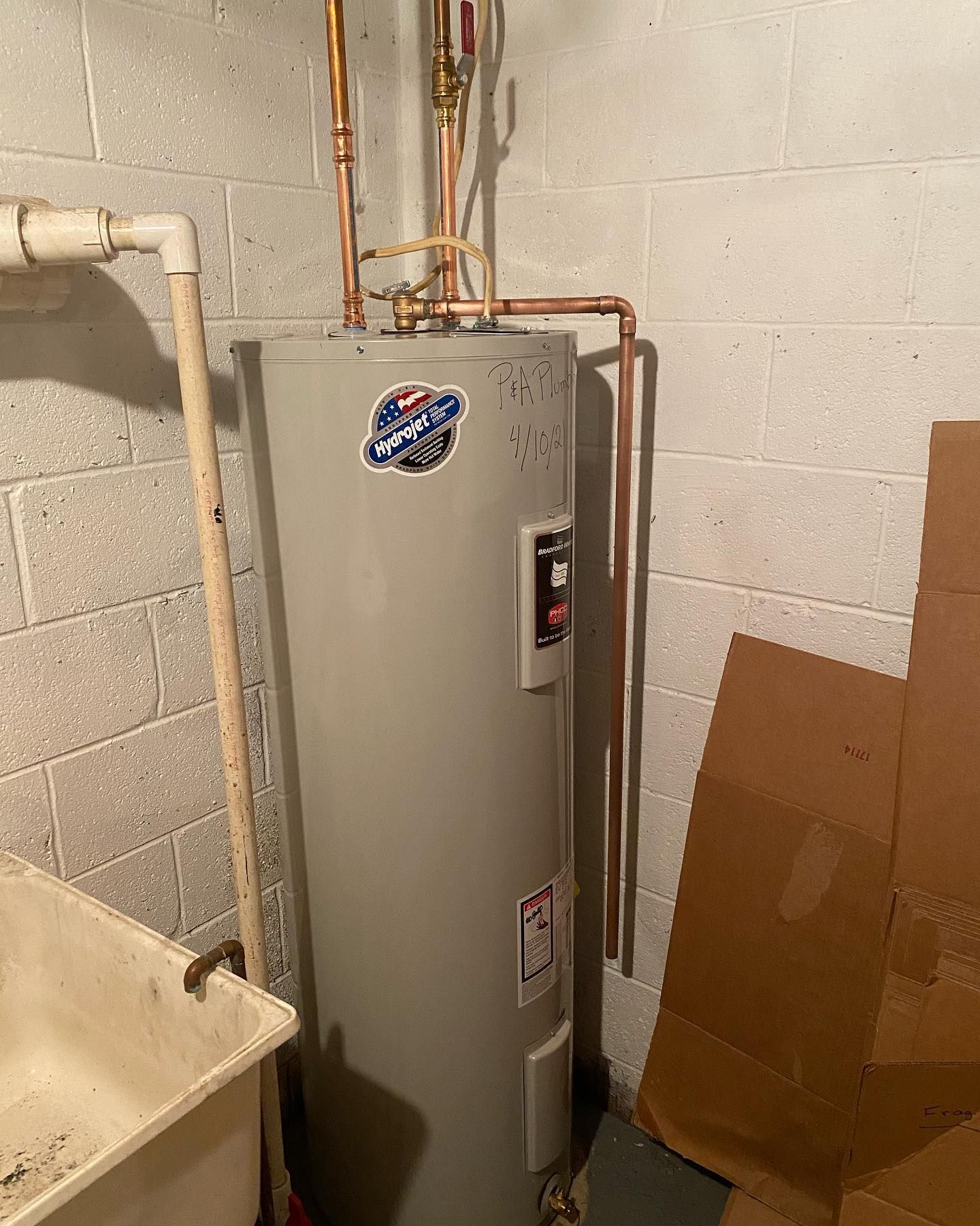 A tall, gray water heater against a cinder block wall with copper pipes, near a utility sink and cardboard.