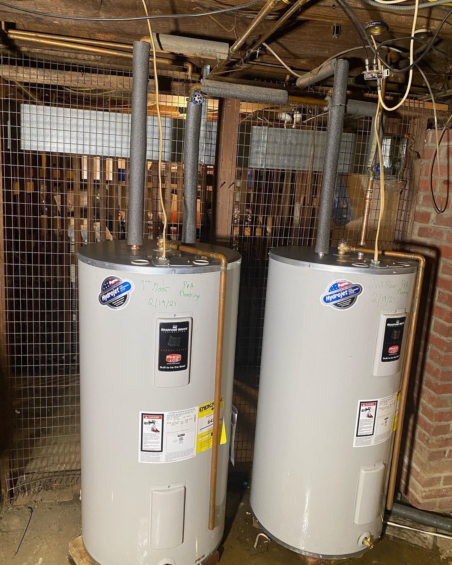 Two tall, silver water heaters in a basement setting, with exposed pipes and wooden beams overhead.