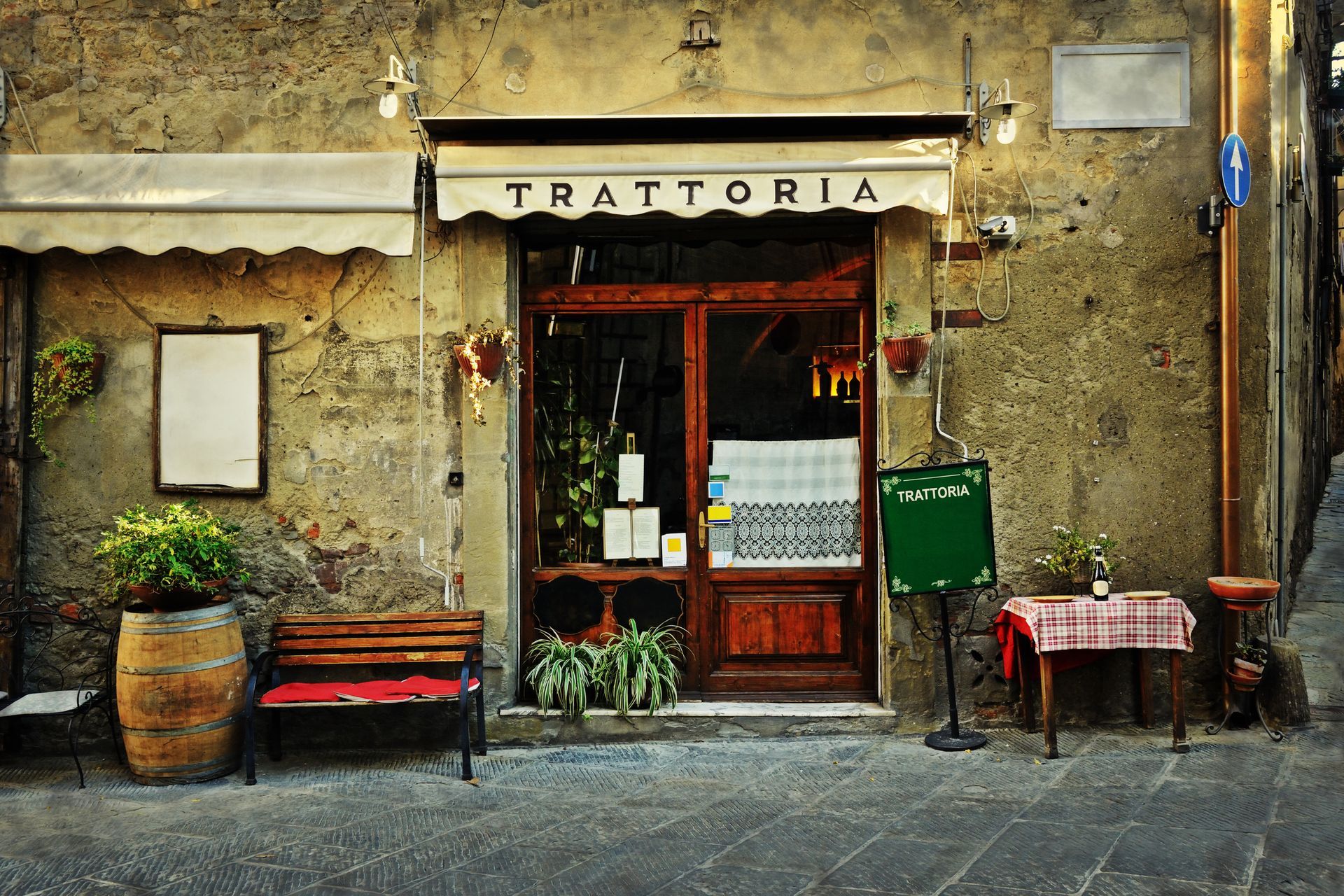 Rustic trattoria storefront with wooden door, potted plants, bench, and red checkered table on a cobblestone street