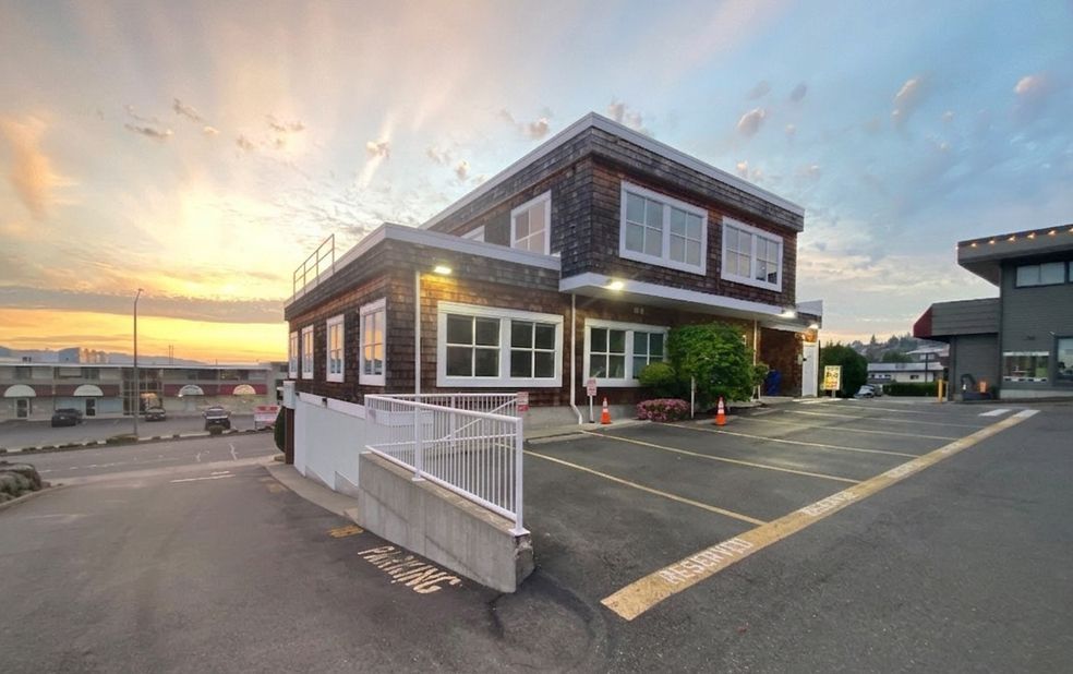 Small waterfront building with white railings at sunset, beside an empty parking lot and road.