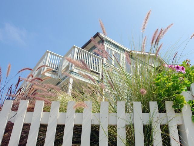 White picket fence with pink flowers and tall grasses in front of a house under a blue sky.