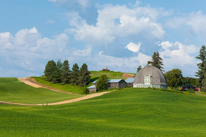 Green rolling fields with a barn, dome-shaped building, and trees under a partly cloudy sky