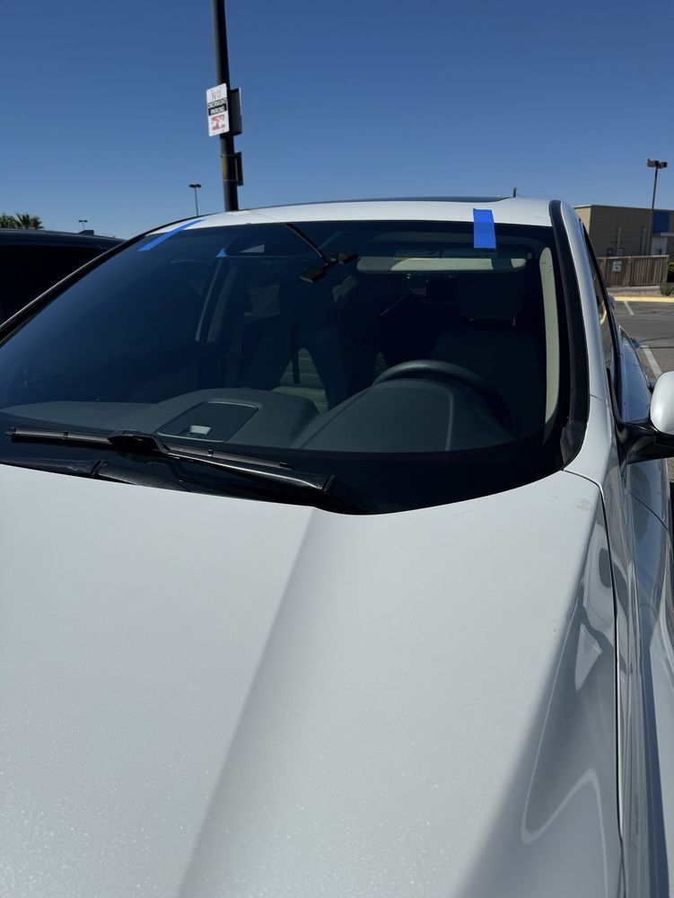 White car with windshield damage, blue tape, parked outdoors on a sunny day.