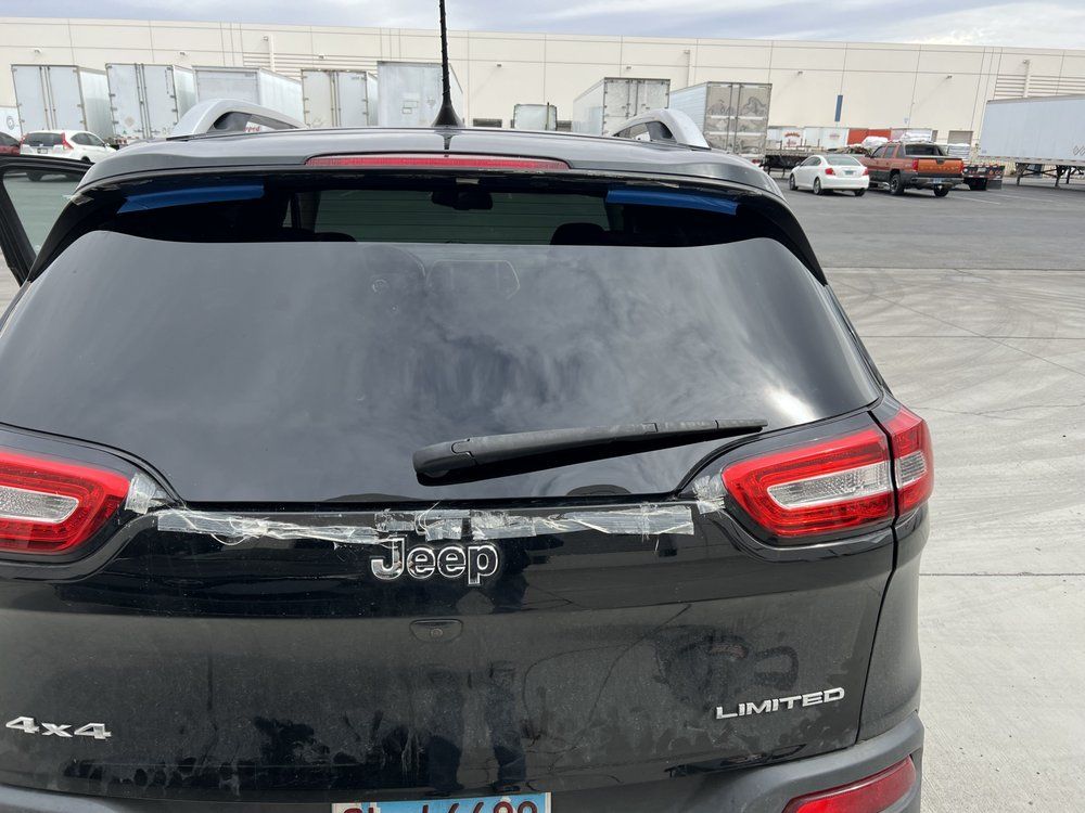 Black Jeep Cherokee with damaged rear window, in an outdoor industrial area.