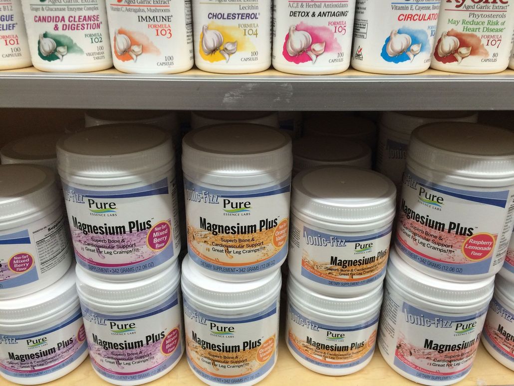 Shelves with jars of Magnesium Plus supplements. White containers with blue and gold labels are stacked.