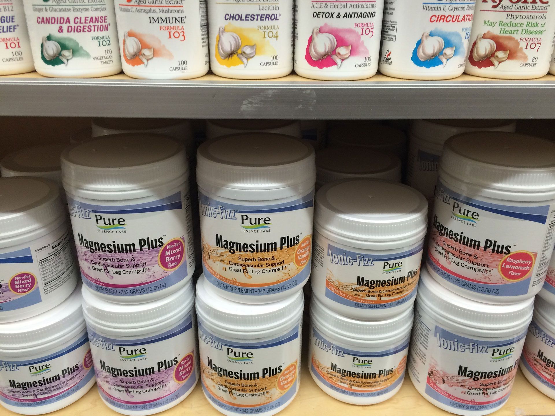 Shelves with jars of Magnesium Plus supplements. White containers with blue and gold labels are stacked.