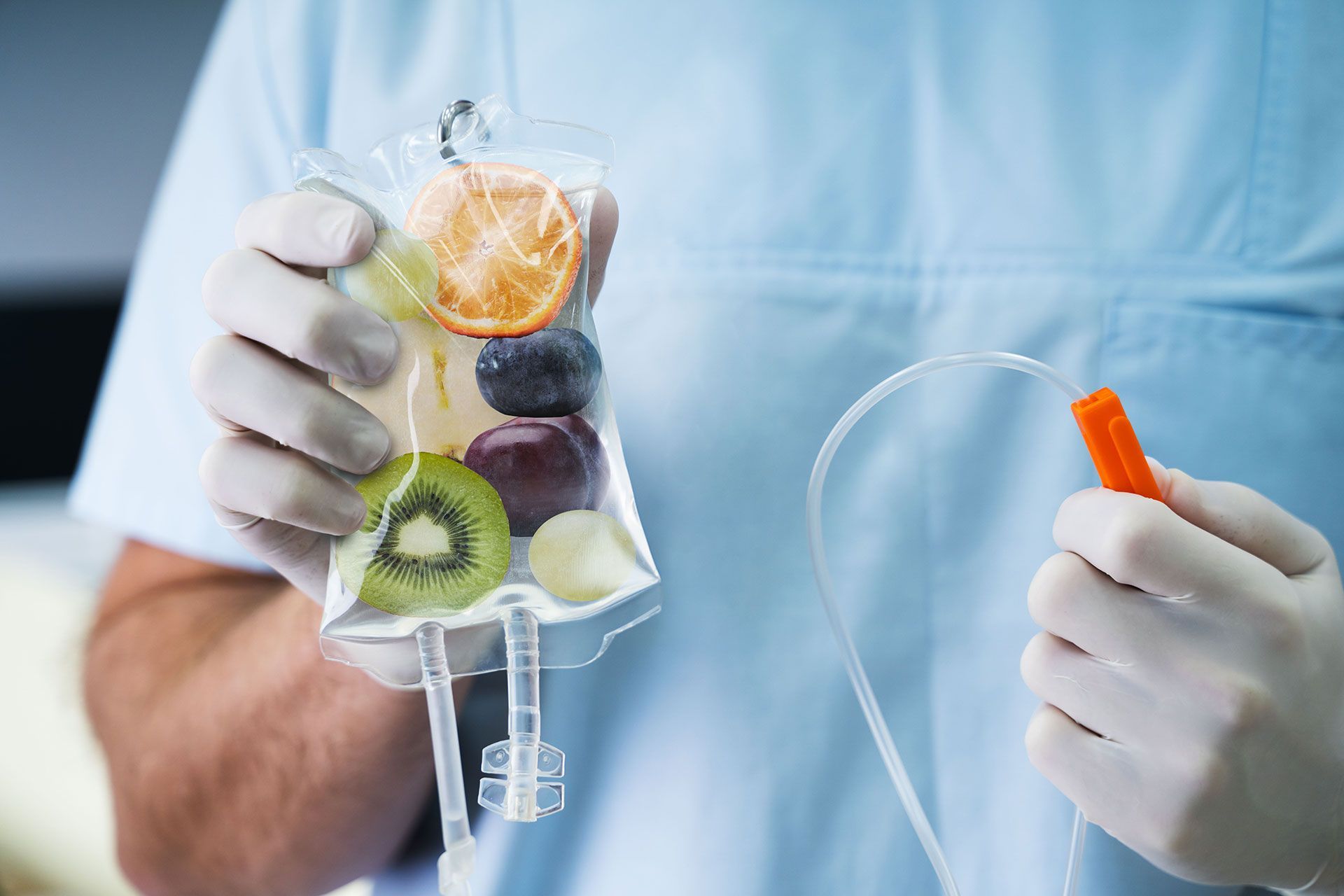 A person wearing gloves holds an IV bag filled with fruit. A clear tube with an orange cap is held in the other hand.
