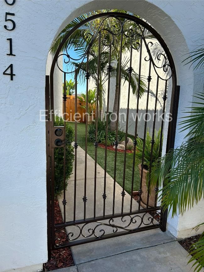 A wrought-iron gate with an arched top and scrollwork opens to a walkway and a green lawn. The gate is set in a white arched entryway.