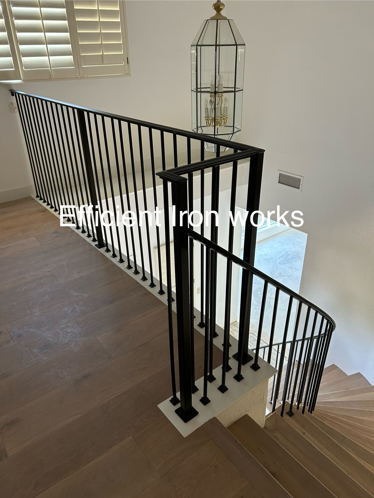 Black metal railing lines a staircase with wooden steps and a light-colored wall; a decorative lamp hangs above.