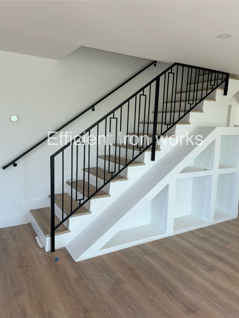 Black iron railing and handrail on a staircase with wooden steps and white wall. Built-in shelves are visible underneath the stairs.