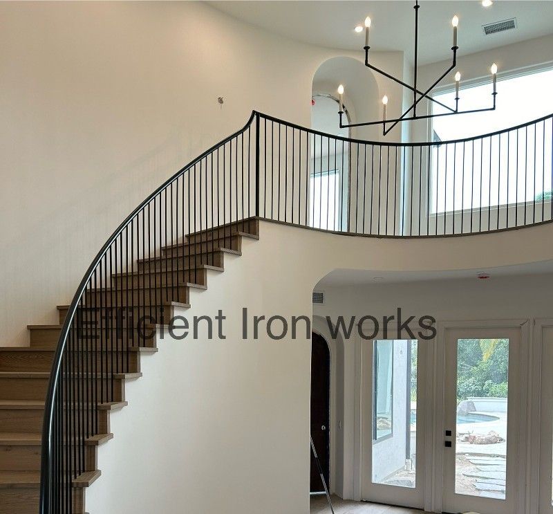A curved staircase with black iron railings, ascending to a balcony. Interior view, light-colored walls and hardwood steps.