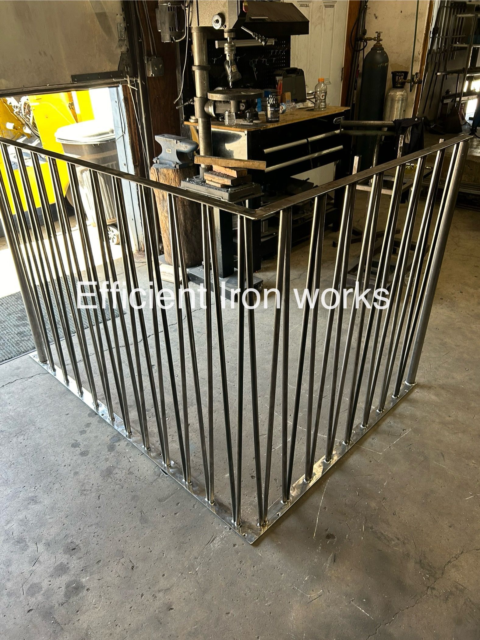 A metal barrier with vertical bars, possibly a railing, in a workshop setting. The metal appears polished, and the floor is concrete.