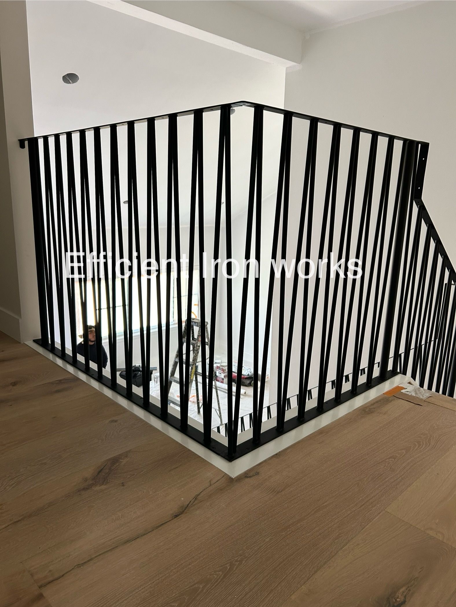 Black metal railing in a corner of a home, providing safety along a staircase. Interior shows a light wood floor and white walls.