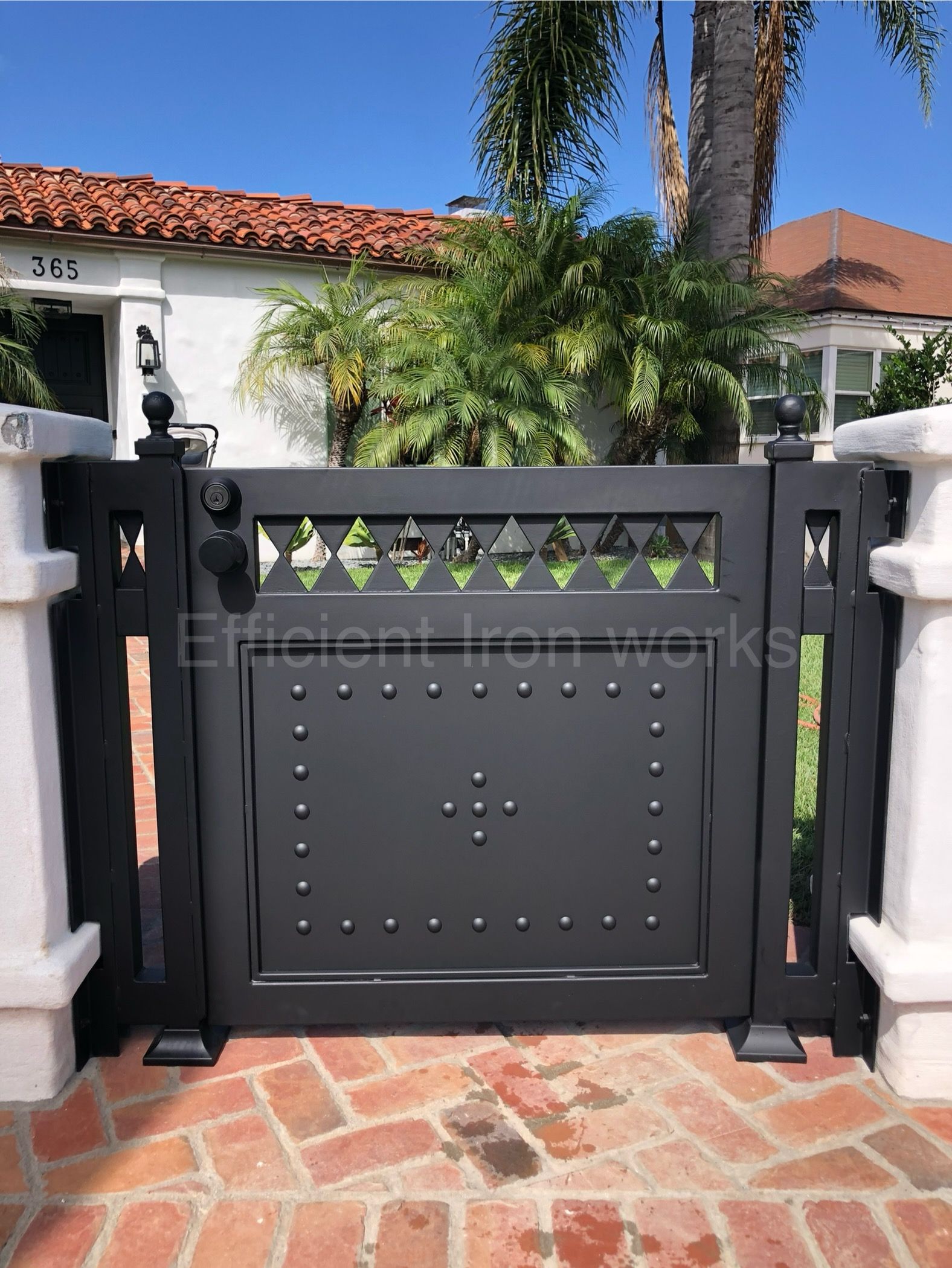 Black wrought iron gate with decorative details set between white pillars, brick path leading to a house with an orange tile roof.