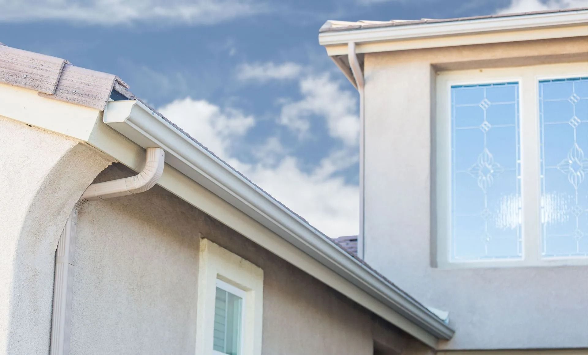 Exterior view of a stucco house with white trim, gutters, and a window with a cloudy blue sky background.