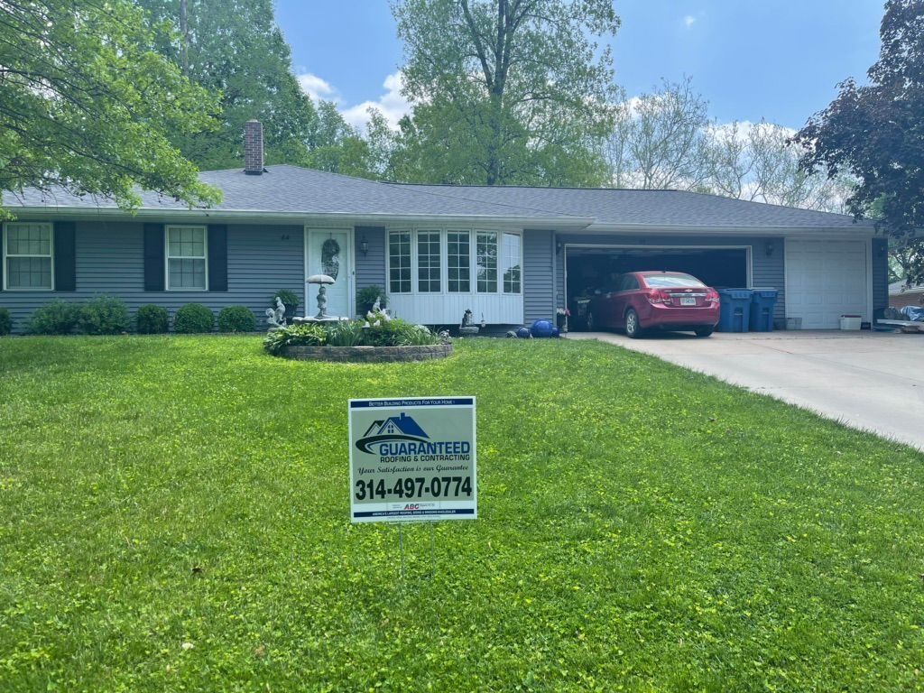 A gray ranch-style house with a new roof, green lawn, car in garage, and a roofing sign.