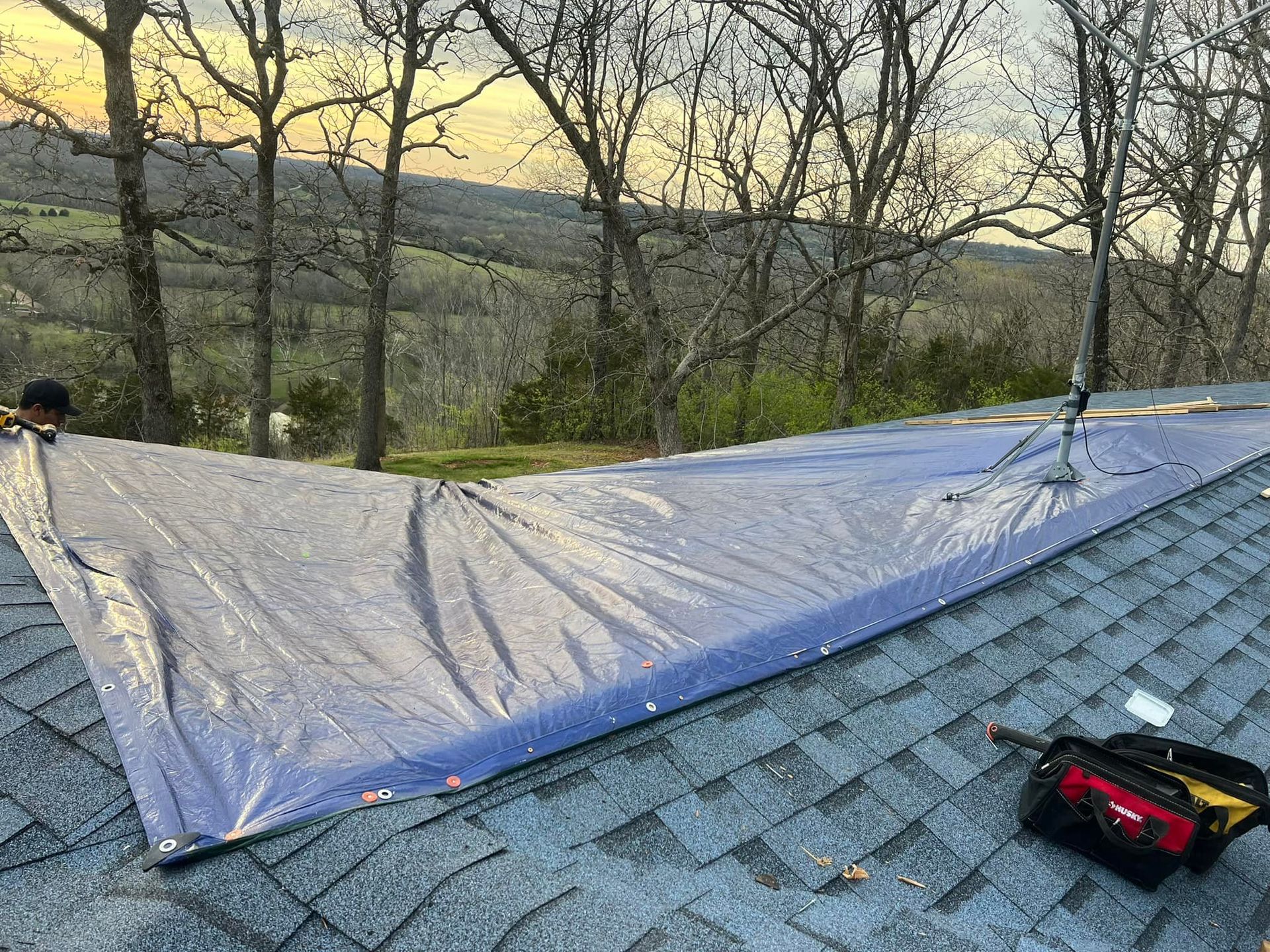 A blue tarp covers a portion of a roof. A person on the roof. Tools in a bag. Trees and hills in the background.