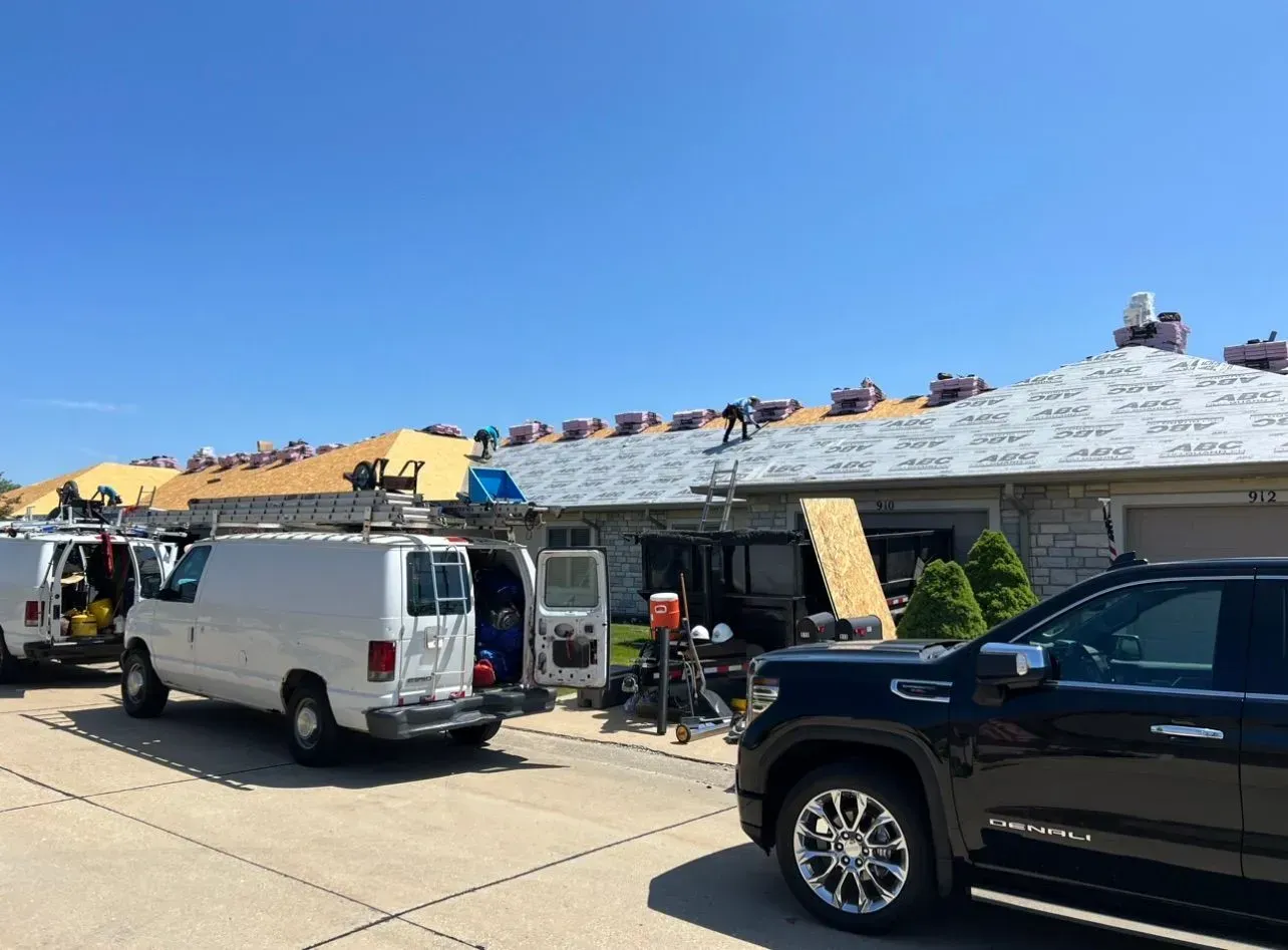 Roofers working on a row of homes with roofing materials. Several vehicles parked on the street. Blue sky.