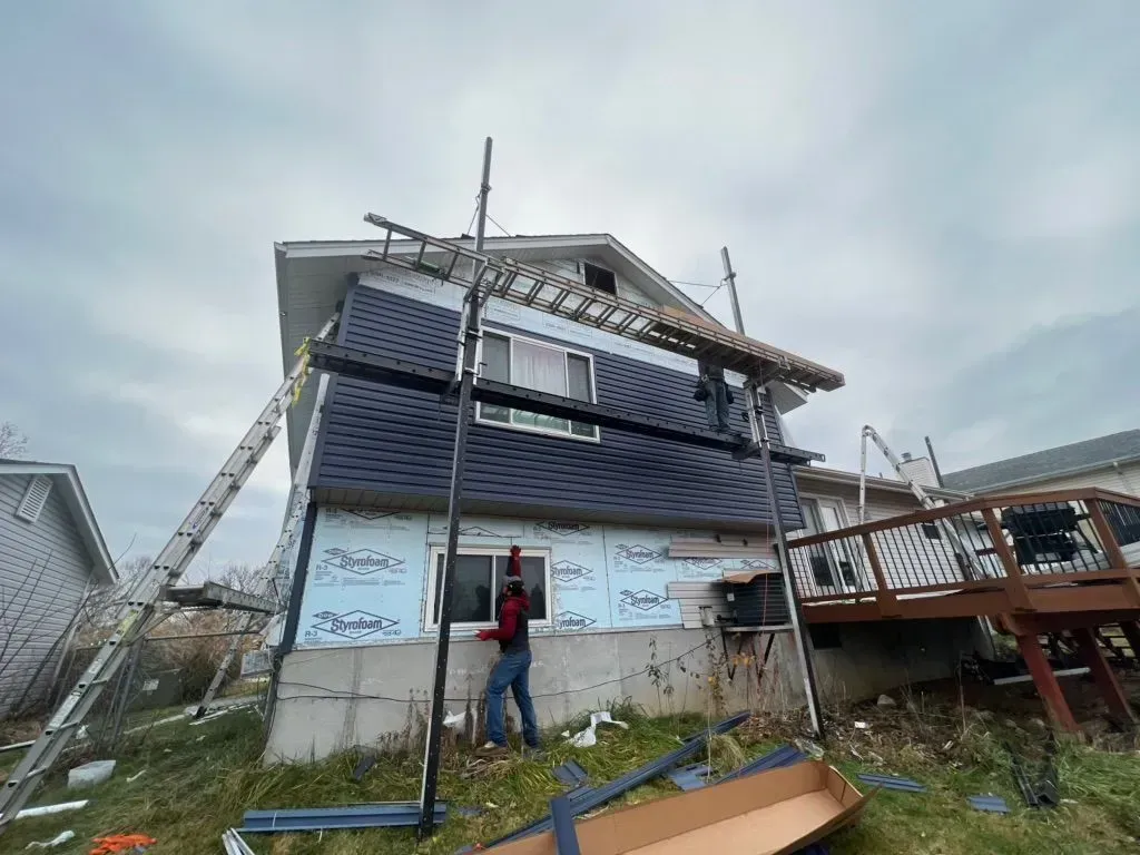 House exterior during siding installation, worker on scaffolding, blue siding, cloudy day.