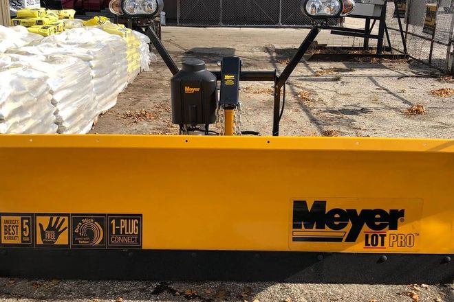 Yellow Meyer snow plow blade with black hydraulic mounts in a yard, stacked materials in the background