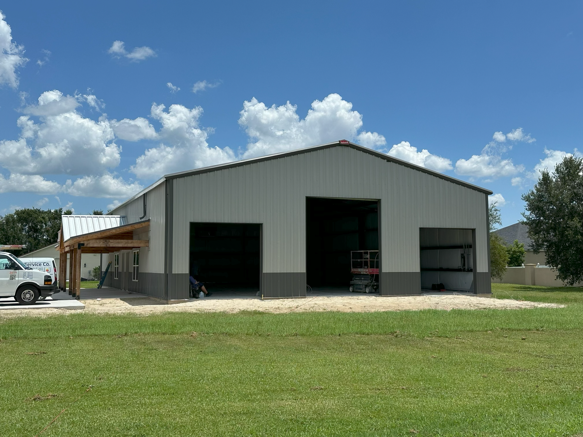 Large gray metal workshop with open bays and a porch, set on a grassy lot under a blue sky.