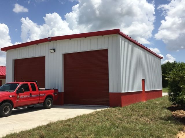 Red metal garage building with two large doors and a red pickup truck parked outside under a cloudy sky