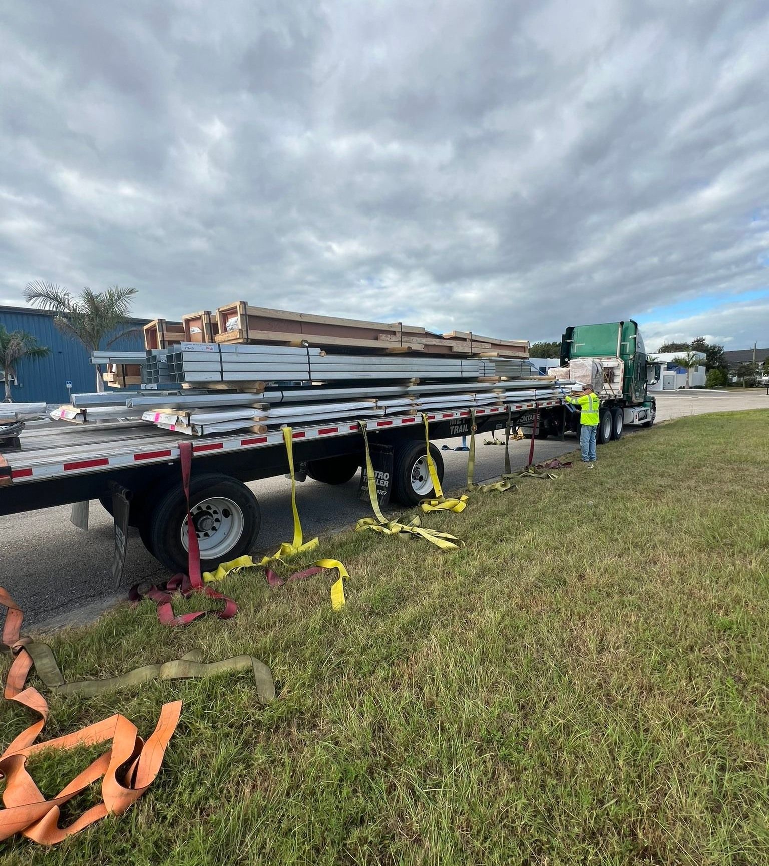 Flatbed truck loaded with lumber at a construction site, with a worker in a neon vest nearby.