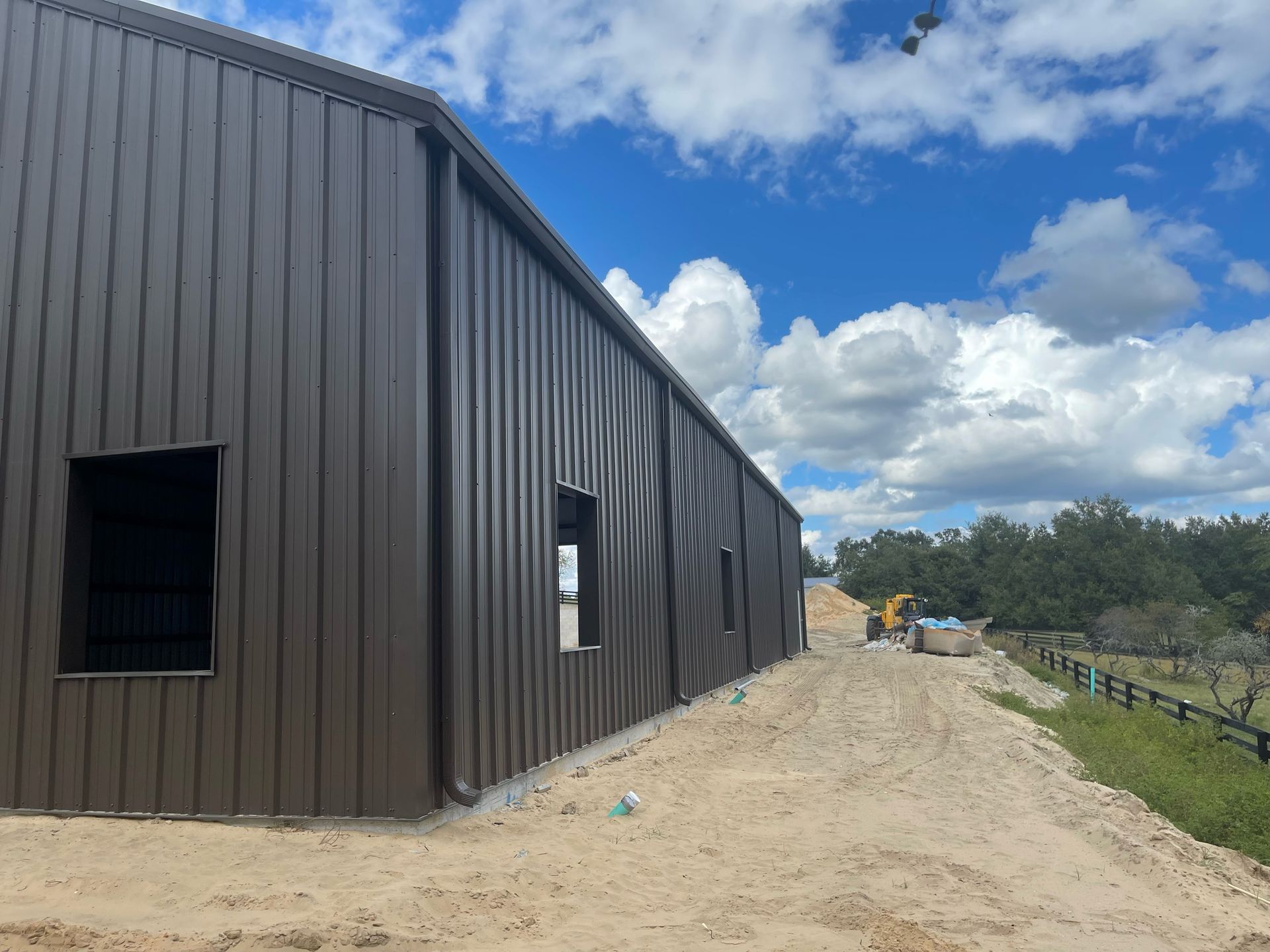 Dark brown metal building beside a dirt road under a blue sky with clouds