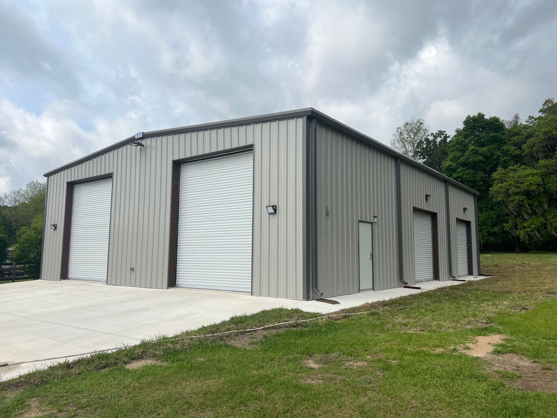 Gray metal storage building with roll-up doors on a concrete pad beside a grassy hillside
