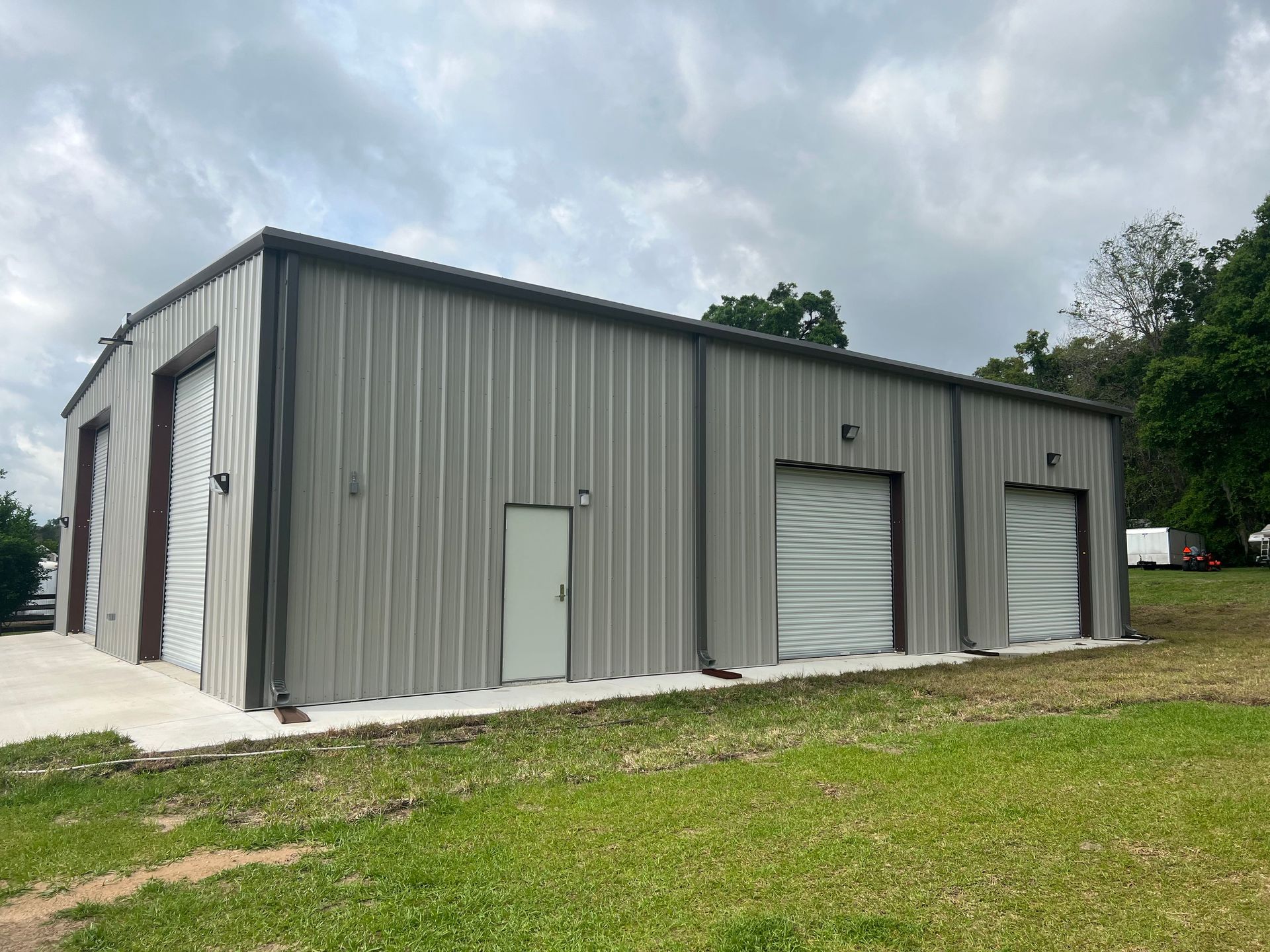 Gray metal storage building with roll-up doors beside a grassy lot under a cloudy sky