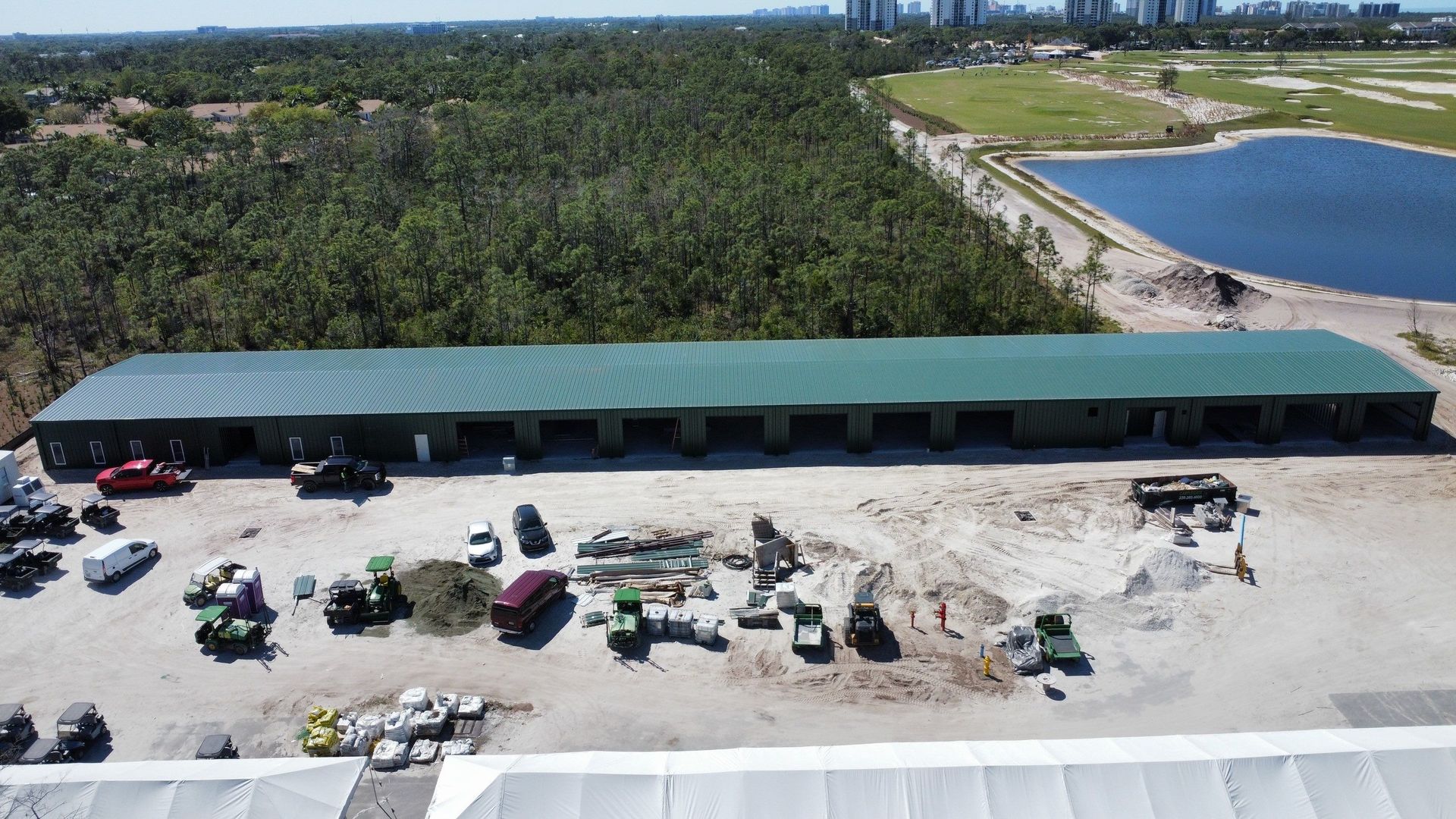 Aerial view of a long green-roofed building beside a sandy waterfront and parked vehicles