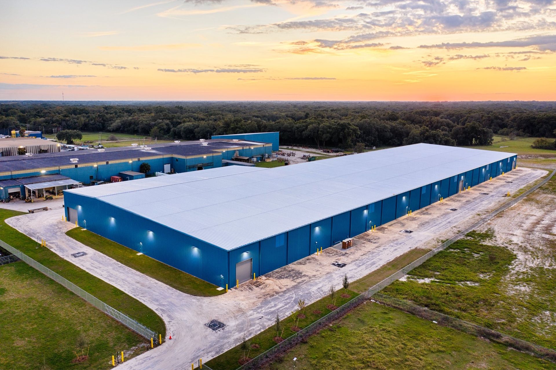 Aerial view of a large blue industrial warehouse at sunset in a rural area.