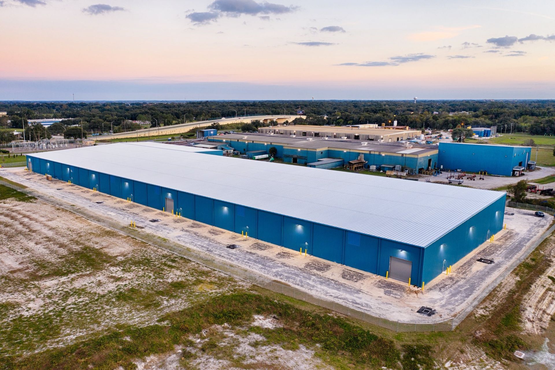 Aerial view of a large blue industrial warehouse complex in a rural area at sunset