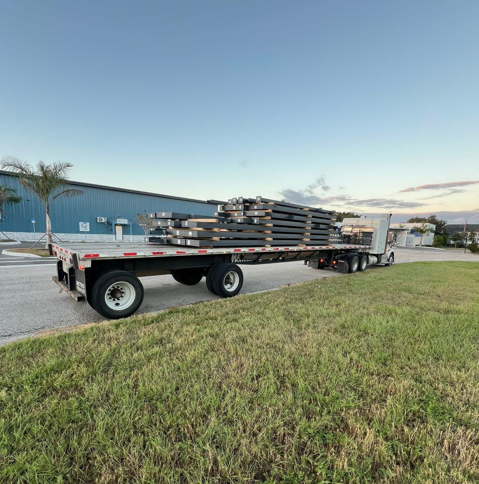 Flatbed trailer loaded with stacked metal beams beside a blue warehouse at dusk