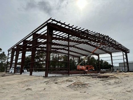 Steel frame building under construction on a dirt lot, with a small orange excavator nearby.