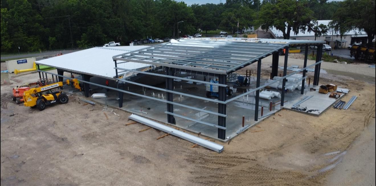 Steel-frame building under construction with a white roof and a yellow loader on a dirt lot