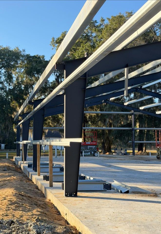 Steel-framed structure under construction on a concrete slab, with white beams and trees in the background