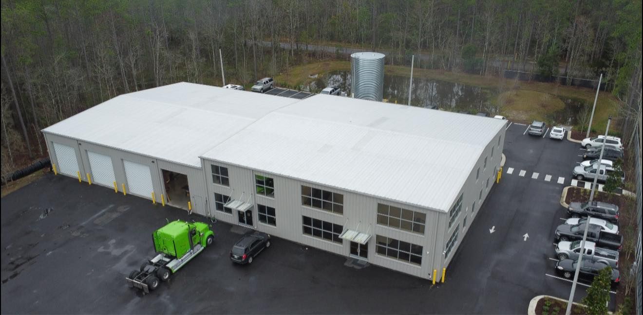 Aerial view of a large white commercial building with a parking lot, trucks, and surrounding trees.