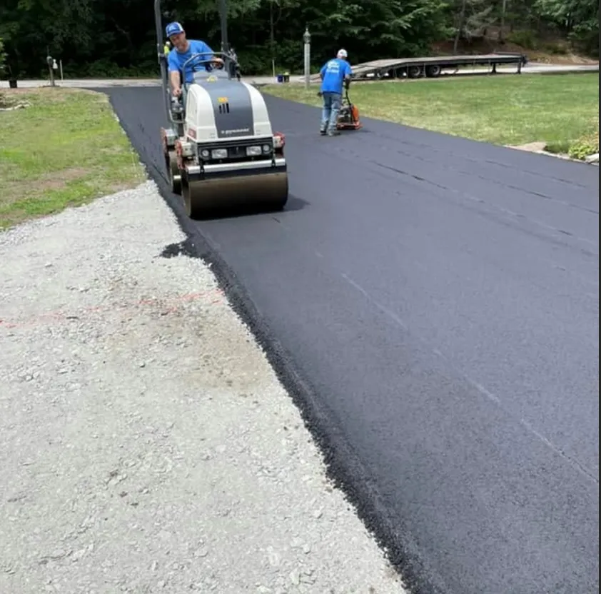 Construction workers use a steamroller and hand-operated compactor to smooth fresh asphalt on a driveway.