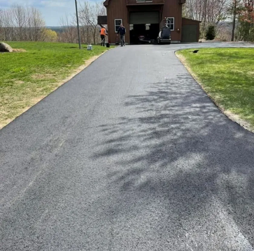 A freshly paved asphalt driveway leading to a brown garage with two people standing near the entrance on a sunny day.