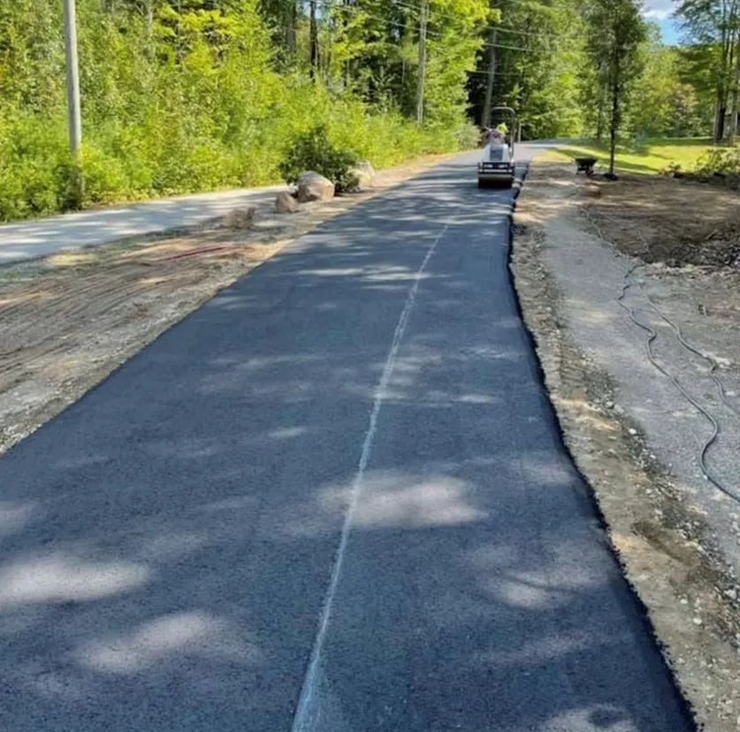 A freshly paved asphalt path winds through a wooded area, with a small construction roller driving in the distance.