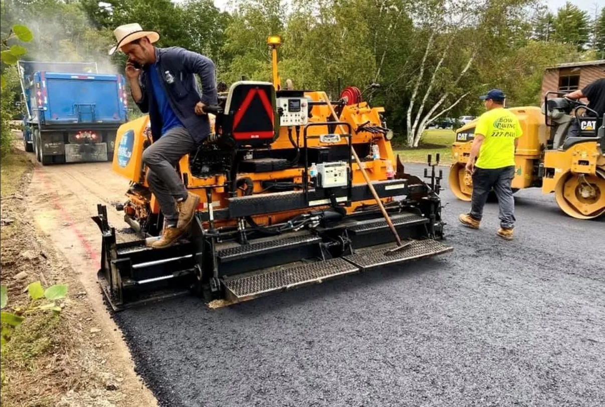 A yellow asphalt paver lays a road surface as a worker in a bright yellow shirt walks nearby.