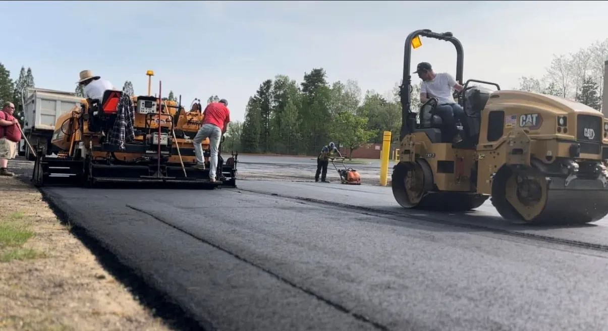 Workers operate a paving machine and a yellow road roller to lay new asphalt on a sunny day.