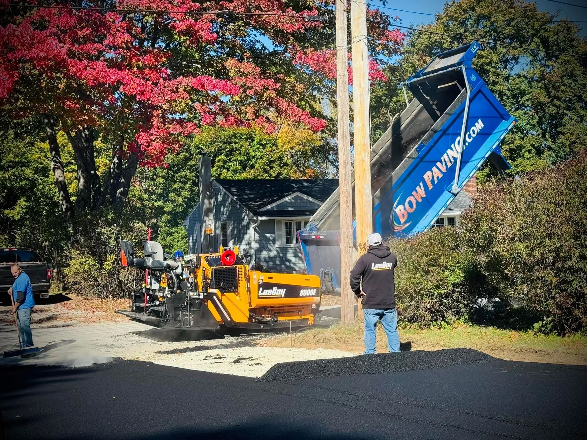 A paving crew works on a driveway with a yellow LeeBoy asphalt paver and a blue dump truck beneath colorful autumn trees.