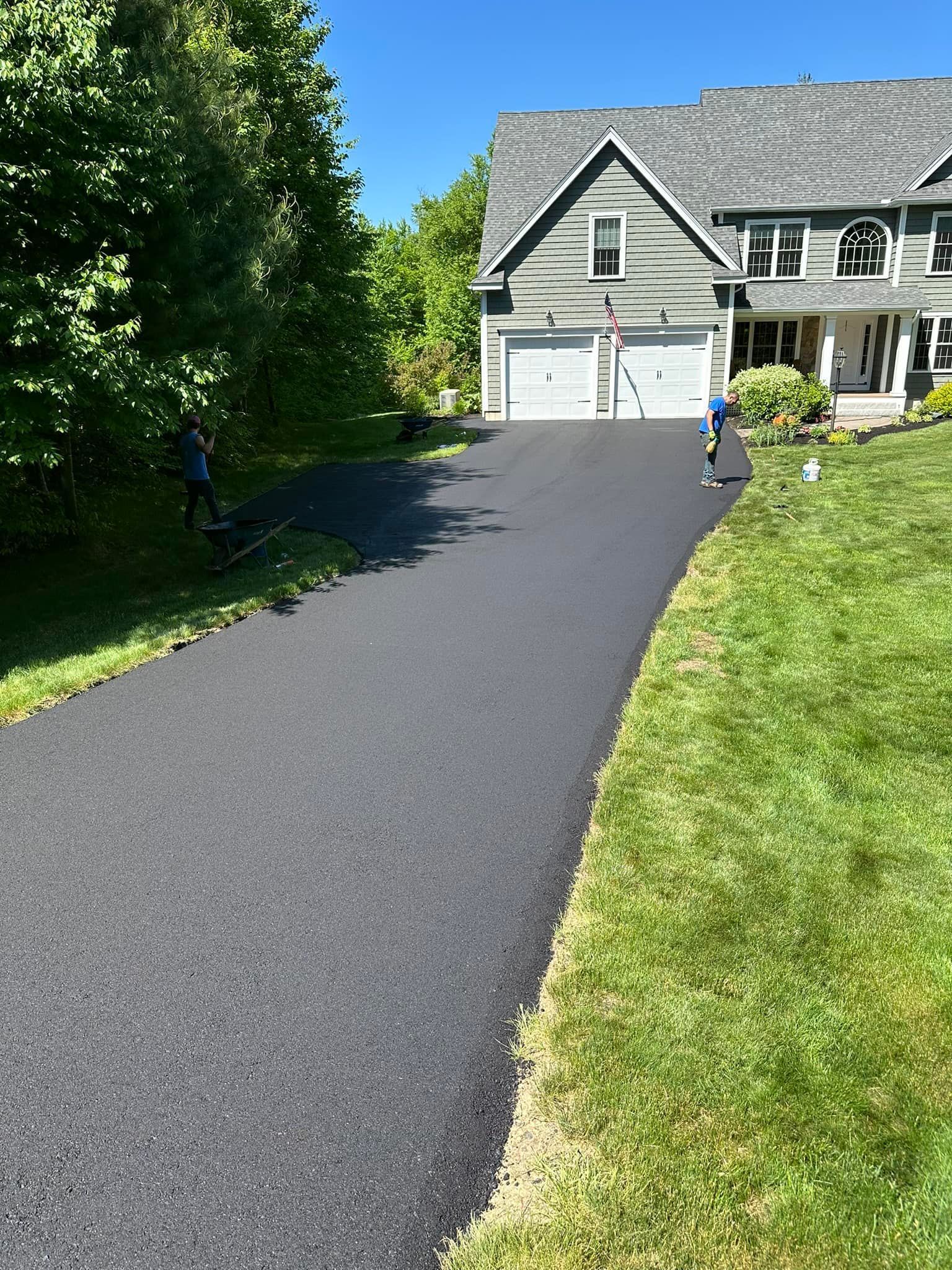 A freshly paved dark asphalt driveway leads up to a two-story house with a grey shingled roof on a sunny day.